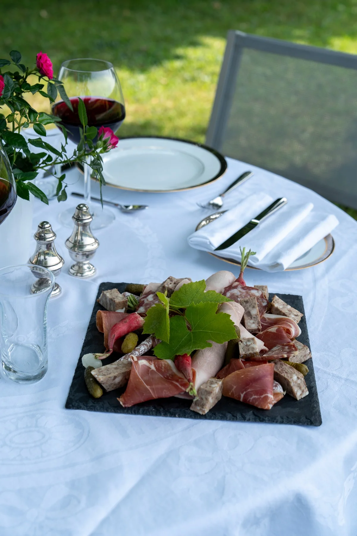 Platter of artisan charcuterie and glasses of red Beaujolais on a flower-adorned table, set in the garden for dinner